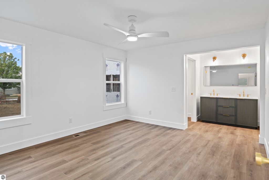 Bright, modern bedroom featuring large windows, wood flooring, and a stylish attached bathroom with dual sinks and contemporary fixtures, showcasing craftsmanship in the new Emerald Hills community, Traverse City.