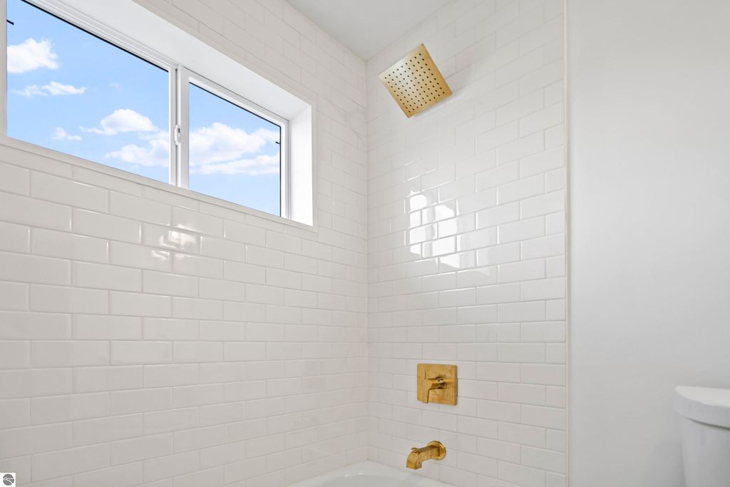 Bright bathroom featuring white subway tile, gold showerhead, and window with blue sky view, showcasing modern design and quality finishes in new Traverse City home.