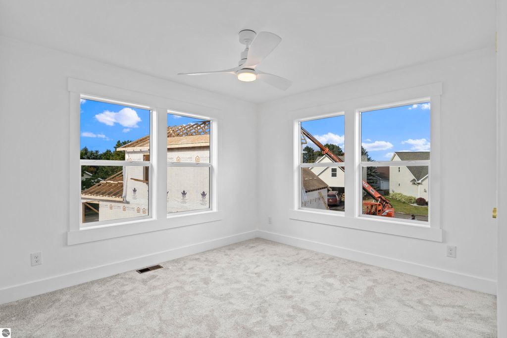 Bright bedroom with large windows showcasing construction views, featuring neutral carpet and modern ceiling fan, reflecting contemporary design in Traverse City home.