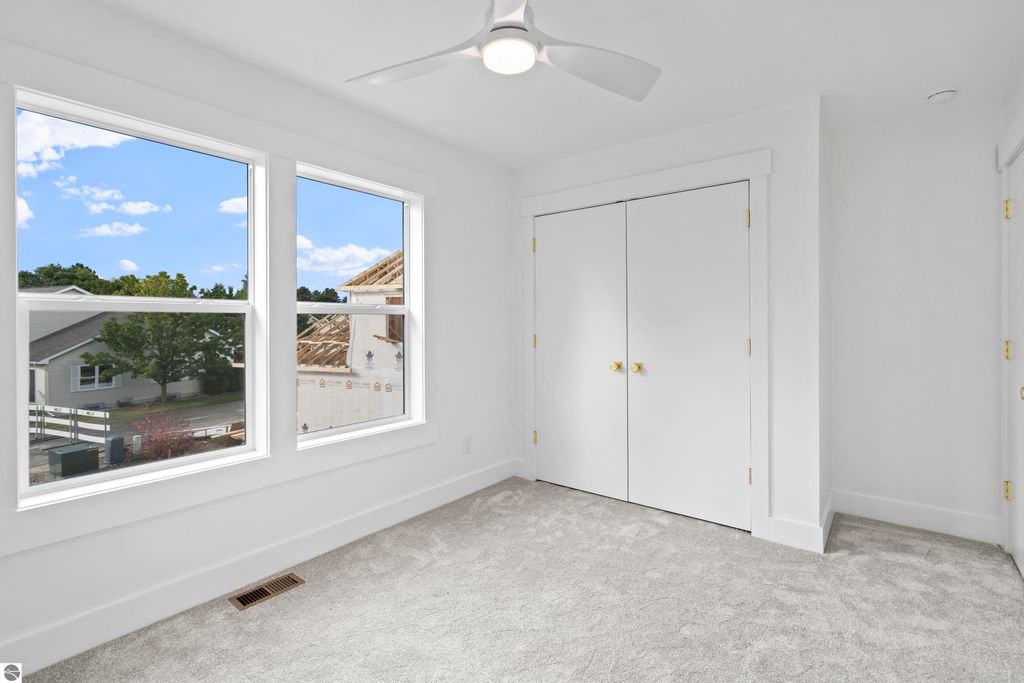 Bright bedroom with large windows showcasing views of the neighborhood, featuring light gray carpet and white walls, complemented by a ceiling fan and double doors to a closet.