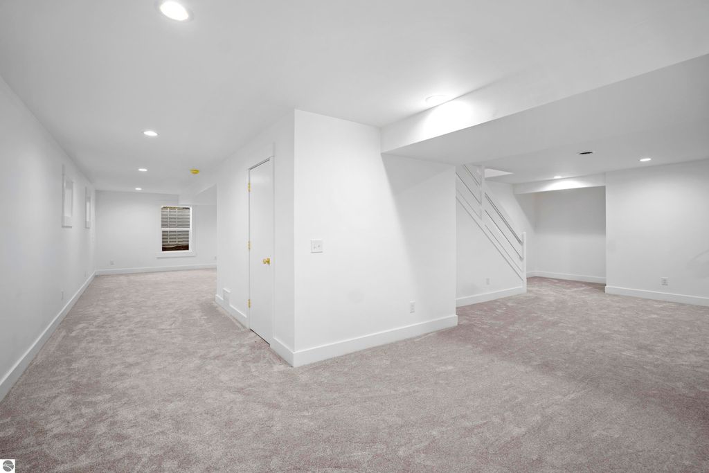 Finished lower level of a modern home in Traverse City, featuring plush carpet, bright white walls, recessed lighting, and a staircase leading to the upper floor, highlighting versatile space ideal for entertainment or home office.
