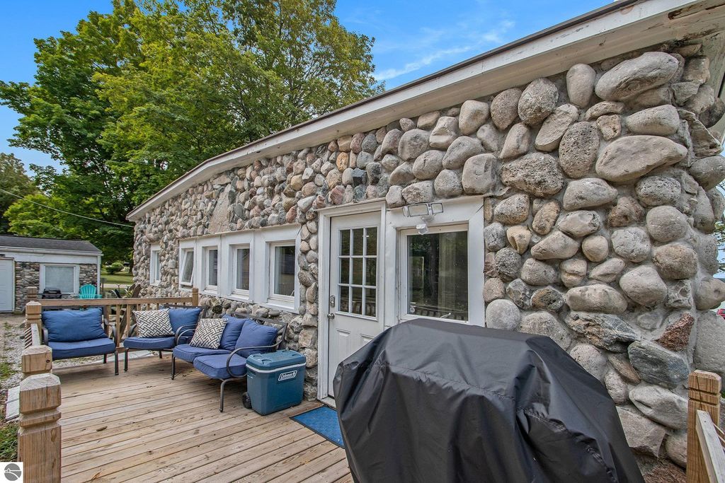 Stone cottage exterior with a rustic design, featuring a pebble stone wall, wooden deck, outdoor seating, and a grill, located near Portage Lake in Onekama, MI, highlighting its rental potential and scenic surroundings.