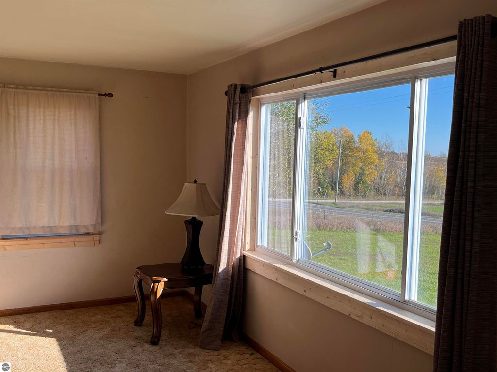Interior view of a cozy room featuring a lamp on a wooden table, large windows with a view of autumn trees outside, and light-colored walls, reflecting the rural charm of the farmhouse at 6034 W Sterling Road, Sterling, MI.