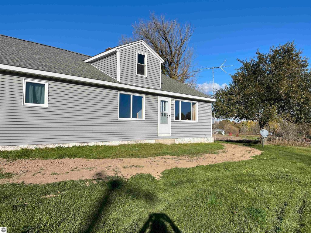 Four-bedroom farmhouse exterior on 2.5 acres in Sterling, MI, featuring gray siding, large windows, and surrounding grassy yard with open views of farmland.