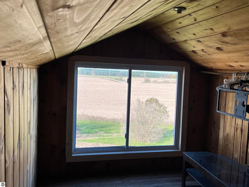 Interior view of a rustic room featuring a large window with a view of open farmland and trees, wooden walls and ceiling, and a small table, illustrating the rural charm of the property at 6034 W Sterling Road, Sterling, MI.
