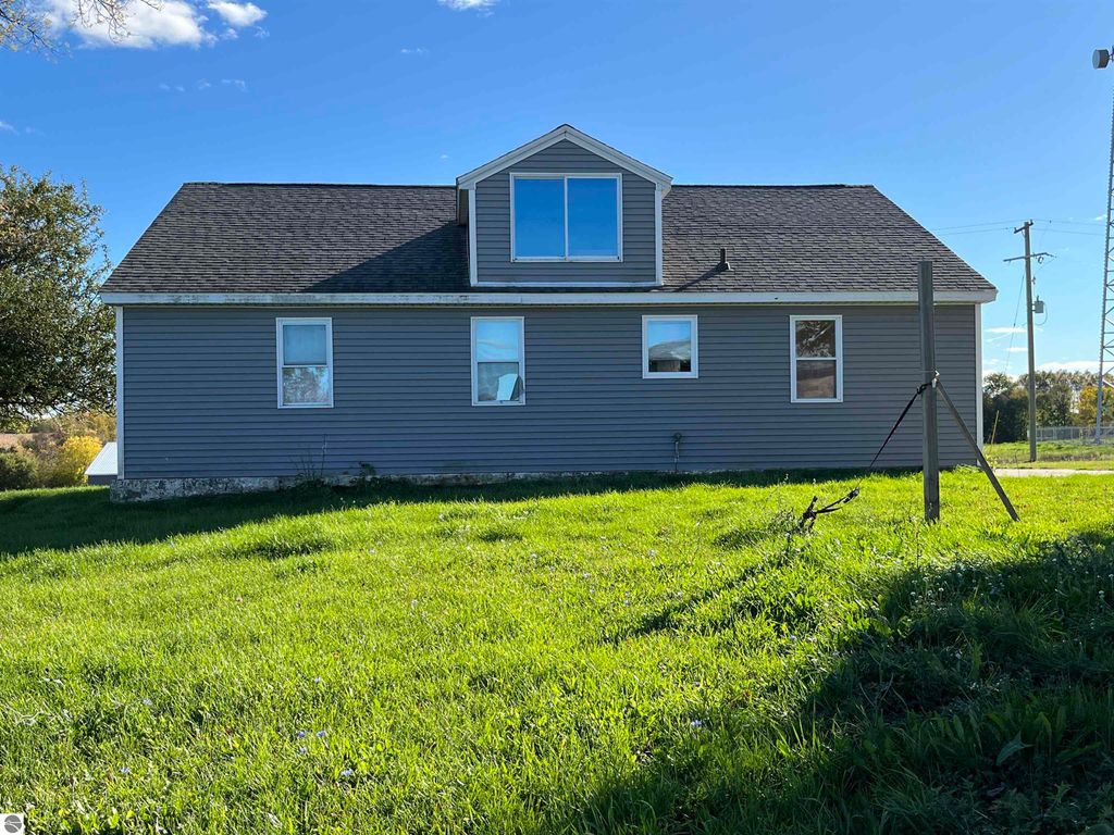 Four-bedroom farmhouse exterior view, gray siding, large windows, situated on 2.4 acres in rural Sterling, MI, surrounded by green grass and open fields.
