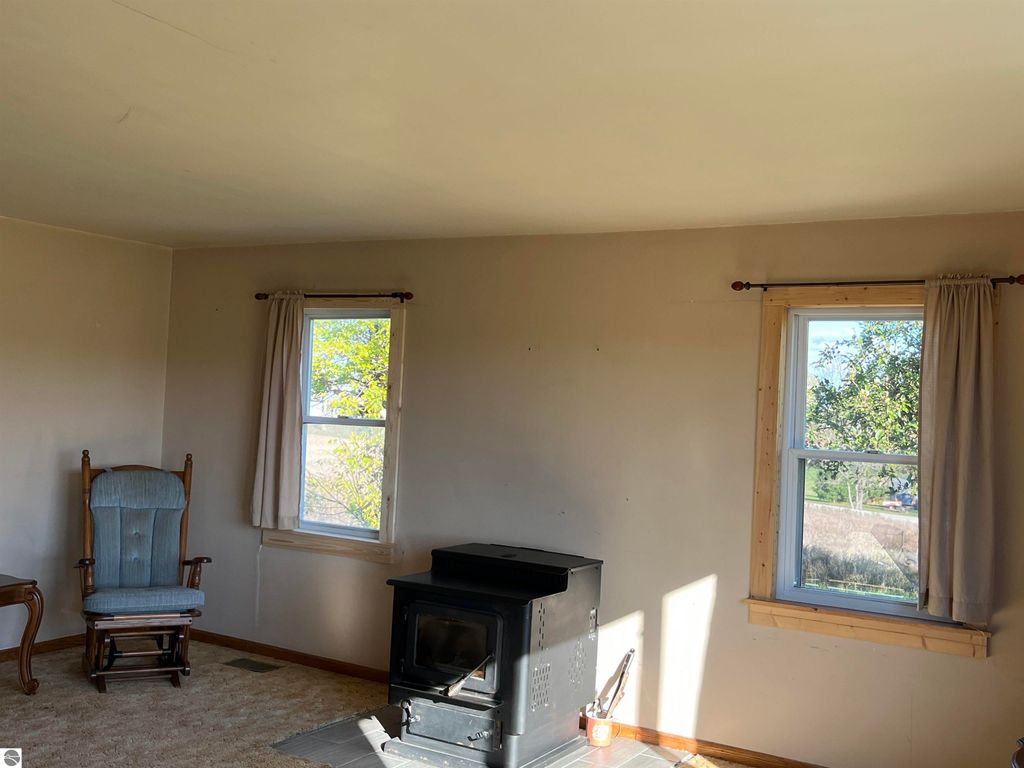 Living room interior of farmhouse at 6034 W Sterling Road, featuring a rocking chair, wood stove, and windows with views of the surrounding landscape.