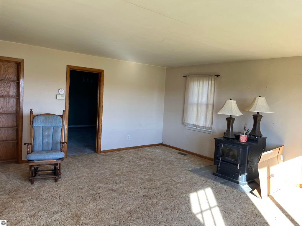 Living room of a four-bedroom farmhouse in Sterling, MI, featuring a rocking chair, a woodstove, and natural light from a window, on carpeted flooring with a neutral color palette.