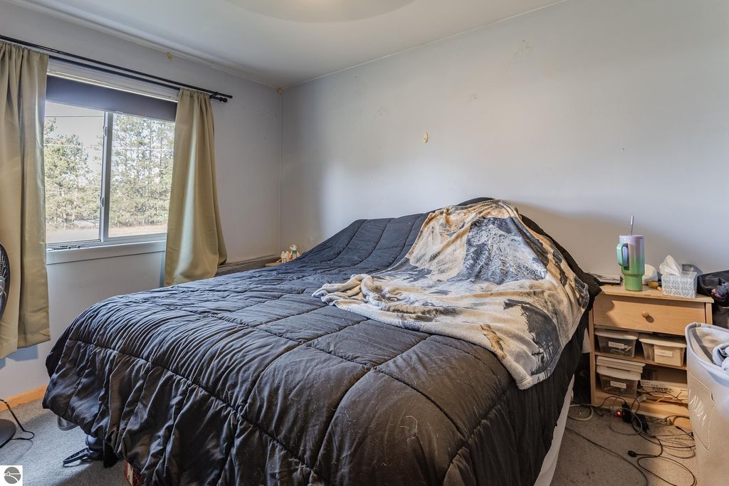 Cozy bedroom featuring a black quilted bedspread, a window with curtains, and a bedside table with storage, reflecting the comfortable living space of the Prescott, MI home for sale at 2265 Michigan Avenue.