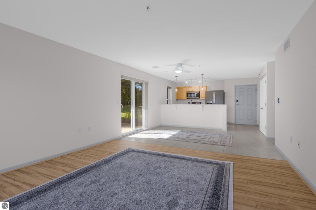First floor living room of a condo in Traverse City, featuring a spacious layout with wooden flooring, a decorative area rug, and a walk-out access to a patio, showcasing modern kitchen appliances in the background.