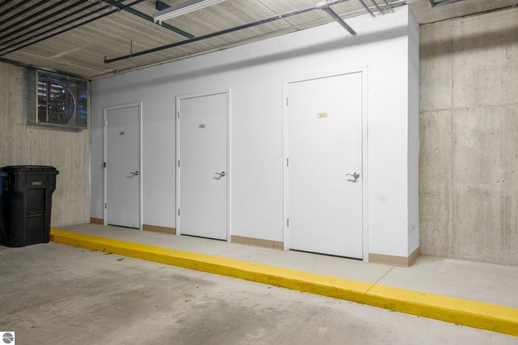 Secure underground parking area featuring three storage units and a trash bin, with a yellow curb for designated parking in a condominium complex in Traverse City, MI.