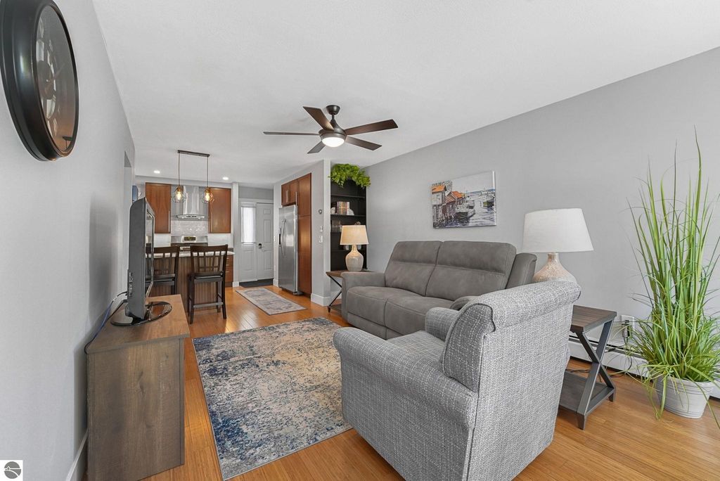Living room of a remodeled townhouse in Traverse City, featuring a gray sofa, wooden dining table, modern kitchen with stainless steel appliances, and decorative wall art, highlighting a bright and open layout.