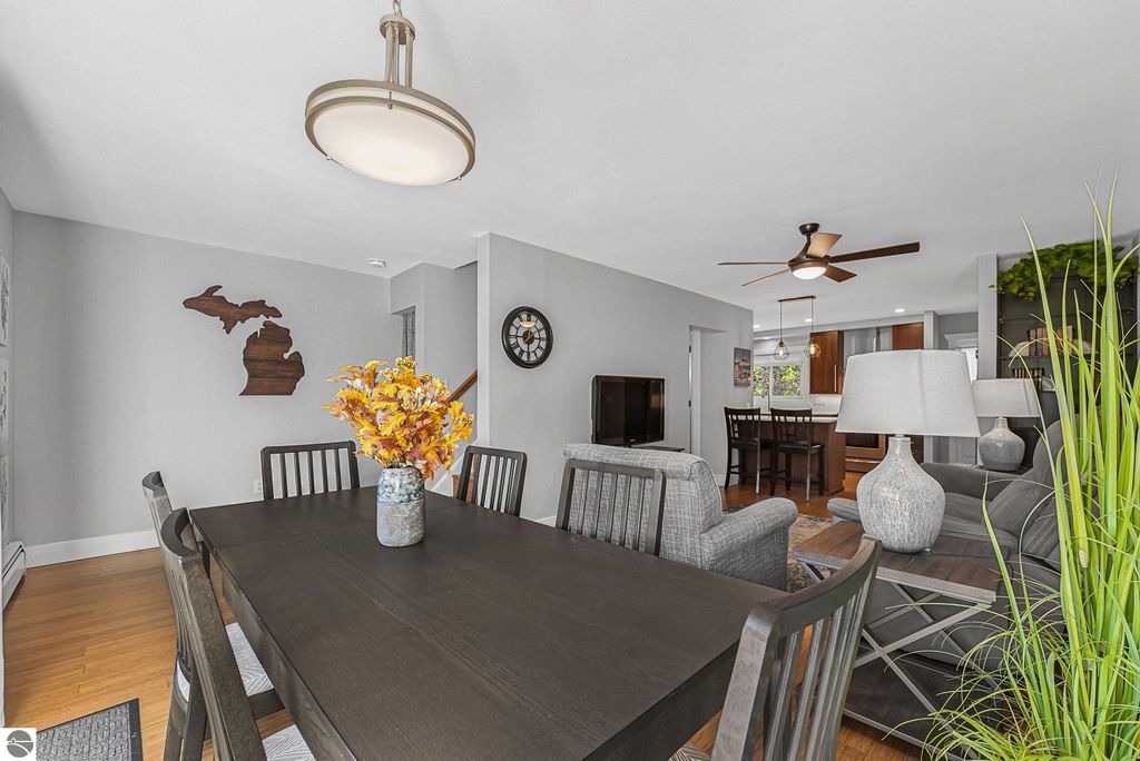 Dining area of a remodeled Traverse City townhouse featuring a dark wooden table with a floral centerpiece, gray upholstered chairs, a Michigan state silhouette wall decor, and a ceiling fan, with an open view of the living space and kitchen beyond.
