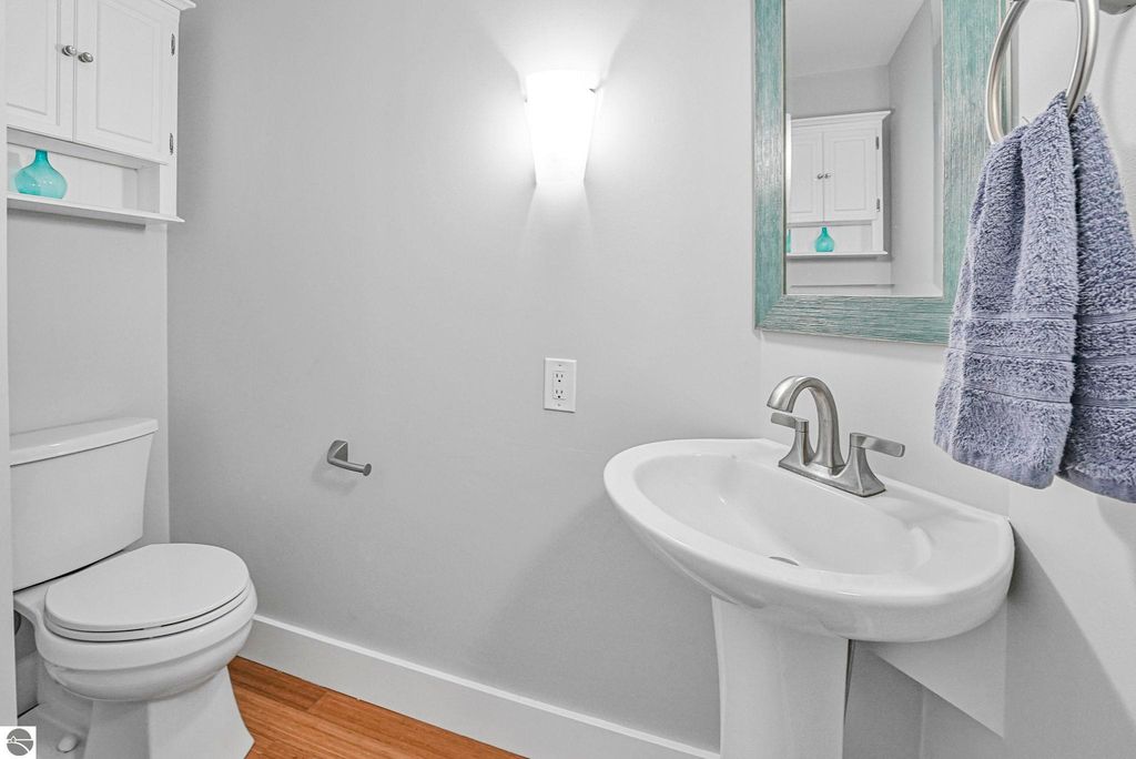 Modern bathroom interior featuring a white pedestal sink with a sleek faucet, a toilet, and a light fixture, complemented by gray walls and wooden flooring, showcasing a towel hanging on a silver ring and decorative shelving.