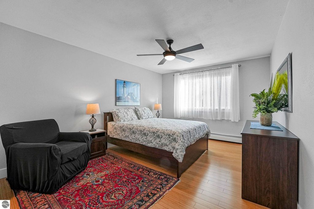 Bright bedroom with a queen bed, decorative bedding, and a cozy armchair, featuring a ceiling fan, hardwood flooring, and large window with sheer curtains, accented by a colorful area rug and modern decor.