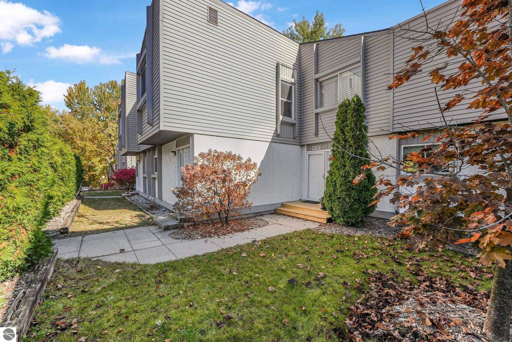 Exterior view of a remodeled townhouse at 10759 S Timberlee Drive #4 in Traverse City, surrounded by greenery and autumn foliage, featuring a small front patio and steps leading to the entrance.