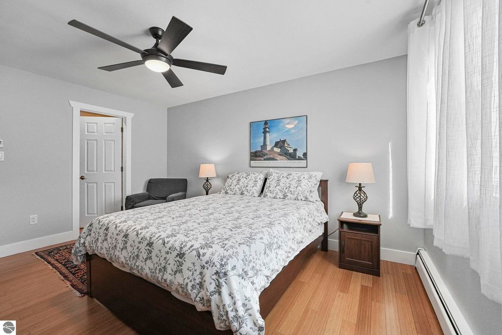 Cozy bedroom in Traverse City townhouse featuring a queen bed with floral bedding, a bedside lamp, and a comfortable chair, accented by a lighthouse painting on the wall and natural light from a window.