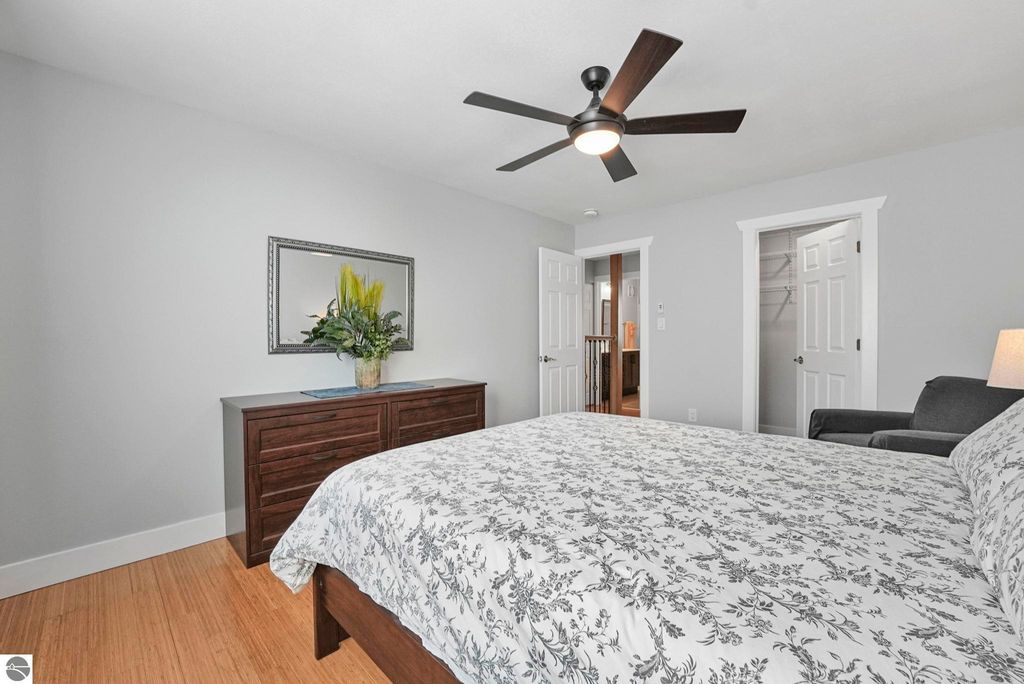 Bright and inviting bedroom in remodeled townhouse, featuring a queen-sized bed with floral bedding, modern ceiling fan, dark wood dresser, and decorative plant, with doors leading to additional rooms and natural light.