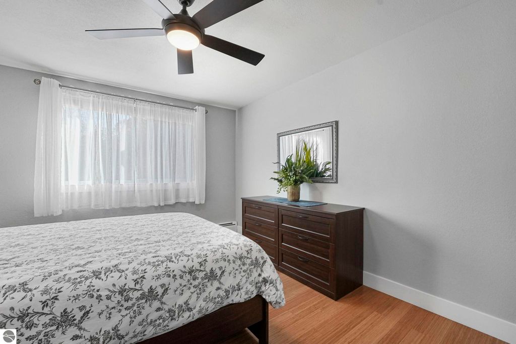 Brightly lit bedroom featuring a bed with floral bedding, a modern dresser, a mirror, and a ceiling fan, showcasing the remodeled interior of the townhouse at 10759 S Timberlee Drive, Traverse City, MI.