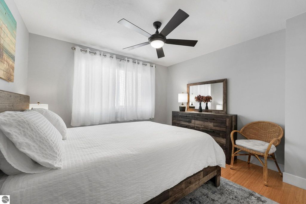 Cozy bedroom featuring a queen-sized bed with white bedding, wooden dresser with mirror, wicker chair, and large windows dressed with sheer curtains, showcasing a bright and inviting atmosphere in the Traverse City townhouse.