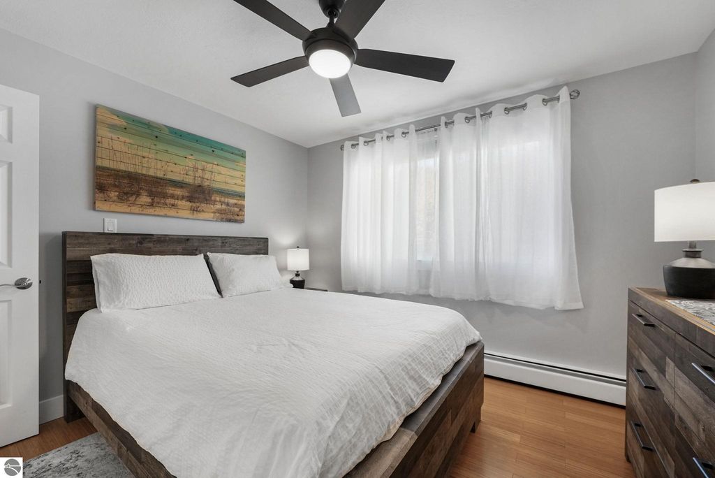 Bright and modern bedroom featuring a queen-sized bed with white bedding, wooden headboard, and a colorful abstract painting on the wall, complemented by natural light from large curtains and a stylish lamp on a nightstand.