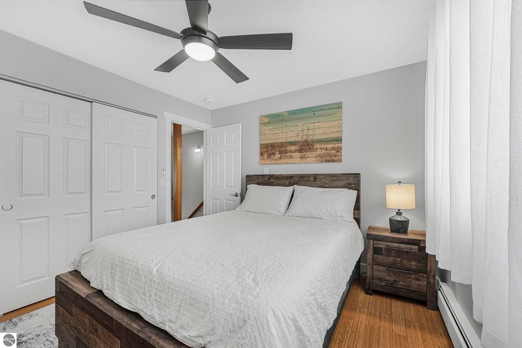 Cozy bedroom featuring a queen-size bed with light bedding, wooden nightstand with lamp, and a colorful wall art piece, complemented by a ceiling fan and natural light from a window, showcasing the inviting atmosphere of the Traverse City townhouse at 10759 S Timberlee Drive.