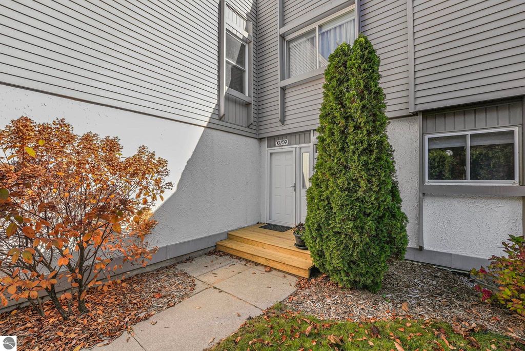 Townhouse entrance at 10759 S Timberlee Drive, featuring a welcoming front door, landscaped shrubs with autumn foliage, and a wooden step leading to the door, highlighting the property’s curb appeal in Traverse City, MI.