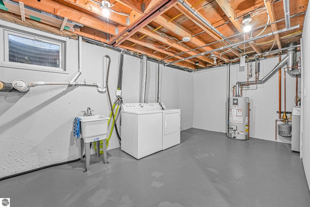 Bright and clean basement laundry area featuring a washing machine, utility sink, water heater, and exposed ceiling with plumbing and electrical fixtures, part of the townhouse listing at 10759 S Timberlee Drive, Traverse City, MI.