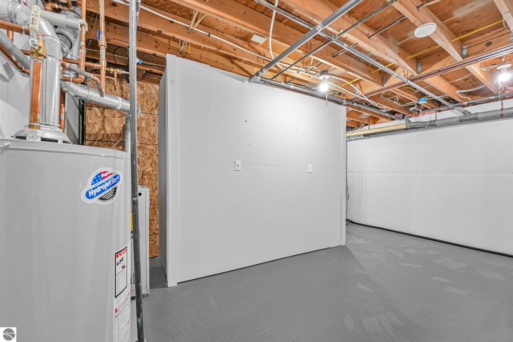 Basement interior of townhouse featuring gray flooring, exposed wooden beams, and utility installations including a water heater, showcasing additional storage space and infrastructure updates.