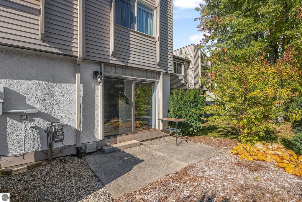 Townhouse patio area with sliding glass door, small table, and landscaped garden featuring shrubs and decorative stones, located at 10759 S Timberlee Drive in Traverse City, MI.