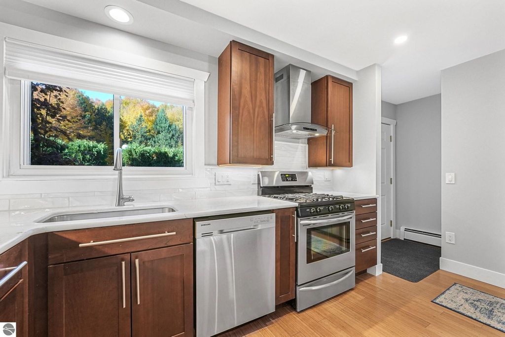 Modern kitchen with stainless steel appliances, quartz countertops, and wooden cabinetry, featuring a window with a view of greenery, part of a remodeled townhouse in Traverse City, MI.