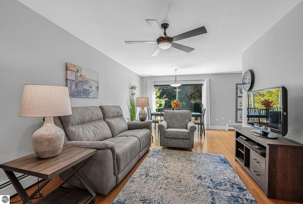 Living room of a remodeled townhouse in Traverse City, featuring a gray color scheme, modern furnishings, a comfortable gray leather sofa, a cozy armchair, a wooden coffee table, and a TV stand, with large windows providing natural light and a view of greenery outside.