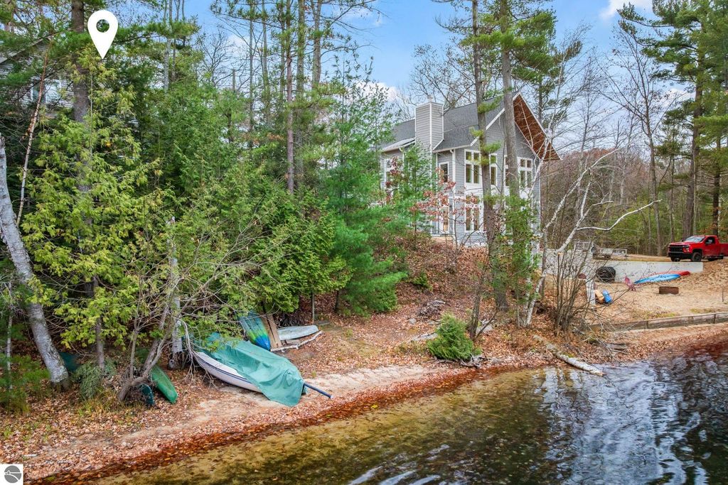 Waterfront view of 4411 Lakeview Trail, Traverse City, MI, featuring sandy shoreline, kayaks, and a modern home surrounded by trees, ideal for relaxed lakeside living.