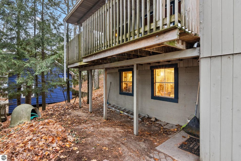 Waterfront home exterior featuring a covered deck overlooking the yard, surrounded by trees and a blue house, with windows illuminated in the lower level.