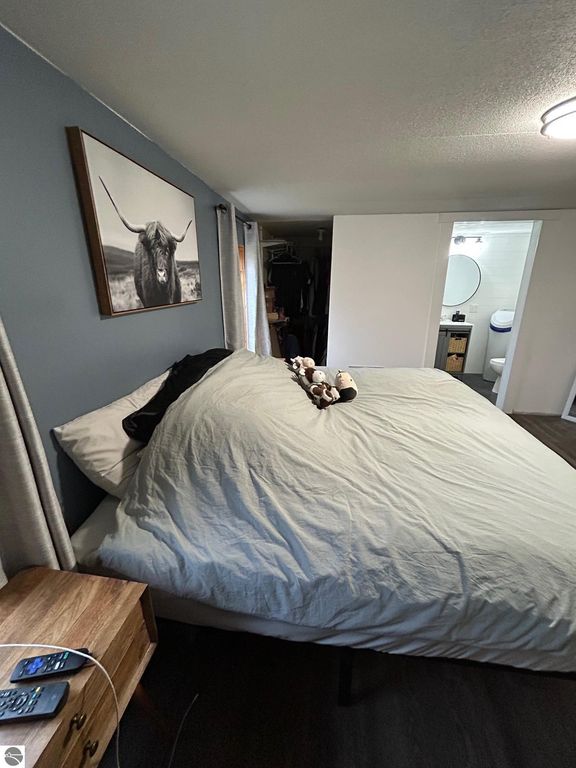 Cozy bedroom featuring a large bed with a light-colored comforter, a wooden nightstand with a phone charger, and a framed black-and-white photo of a longhorn steer on the wall, with an adjoining bathroom visible in the background.