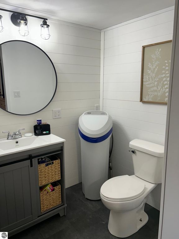 Updated bathroom with a circular mirror, modern lighting, and a sleek vanity, featuring a toilet and a unique trash bin, showcasing contemporary design elements in a home setting.