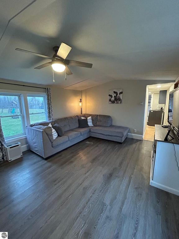 Cozy living room featuring a gray sectional sofa, ceiling fan, and large windows with views of the yard, showcasing updated flooring and modern decor in a residential property for sale at 3780 E North County Line Road, St Louis, MI.