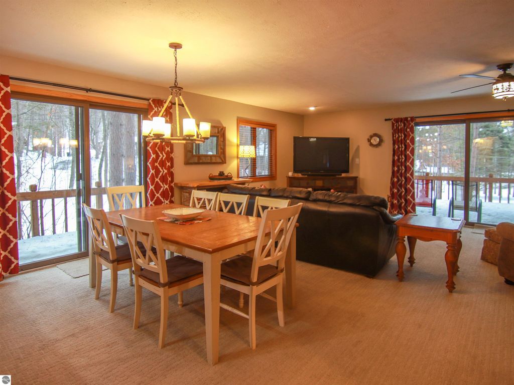 Cozy dining area with wooden table and chairs, living room featuring a leather couch and television, large windows overlooking snowy outdoor scenery, and decorative accents in a Crystal Mountain cottage setting.