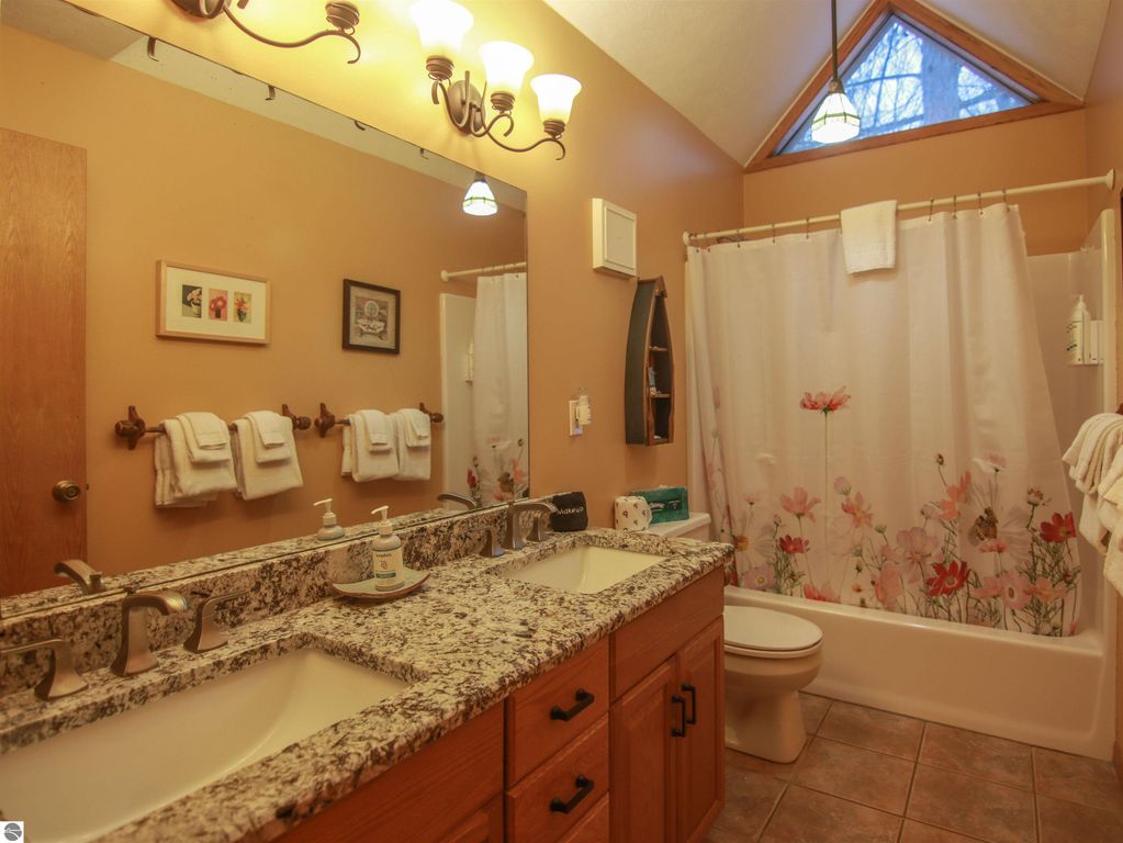 Spacious bathroom featuring dual sinks, granite countertop, floral shower curtain, and natural light from a triangular window, highlighting a cozy cottage atmosphere in Thompsonville, MI.