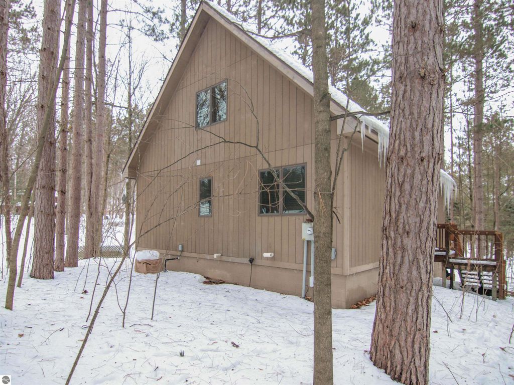 Cozy cottage at 7864 Chippewa Drive, surrounded by tall pines and snow, showcasing the wooden exterior and nearby deck, ideal for winter getaways in Thompsonville, MI.
