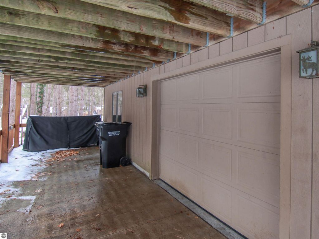 Covered carport area with a garage door, trash bin, and a view of surrounding trees, highlighting the outdoor space of the cottage at 7864 Chippewa Drive, Thompsonville, MI.