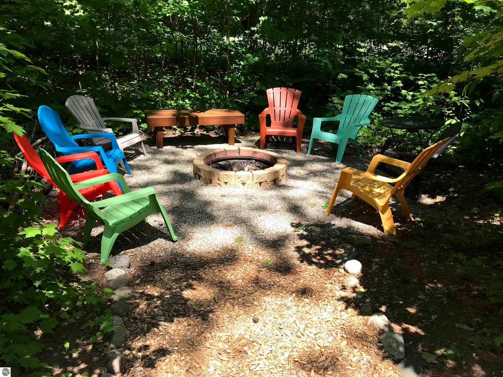 Colorful Adirondack chairs surrounding a stone fire pit in a wooded outdoor setting, ideal for gatherings near the Crystal Mountain cottage at 7864 Chippewa Drive, Thompsonville, MI.