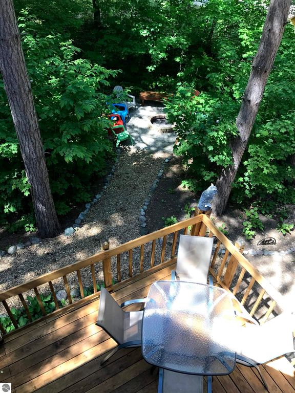 Wooden deck view overlooking a landscaped backyard with a gravel path, fire pit, and colorful outdoor chairs surrounded by lush greenery, highlighting the tranquil outdoor space of the Crystal Mountain cottage at 7864 Chippewa Drive.