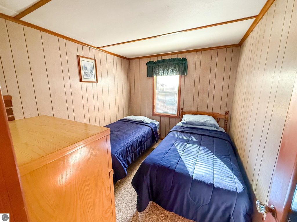 Cozy bedroom featuring two twin beds with blue bedding, wooden furniture, and warm wood-paneled walls, ideal for a cottage getaway in Prudenville, MI.