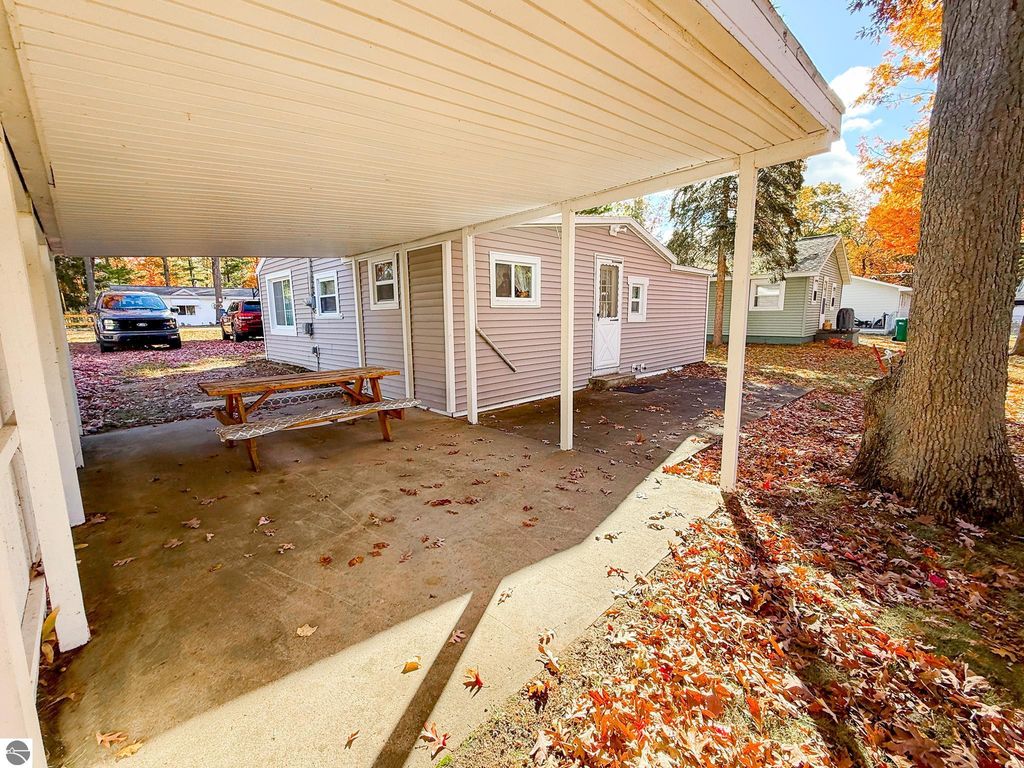 Cottage exterior at 133 Maple Bluffs Avenue, Prudenville, MI, featuring a covered carport, picnic table, and autumn leaves, showcasing the cozy outdoor space ideal for relaxation and recreation.