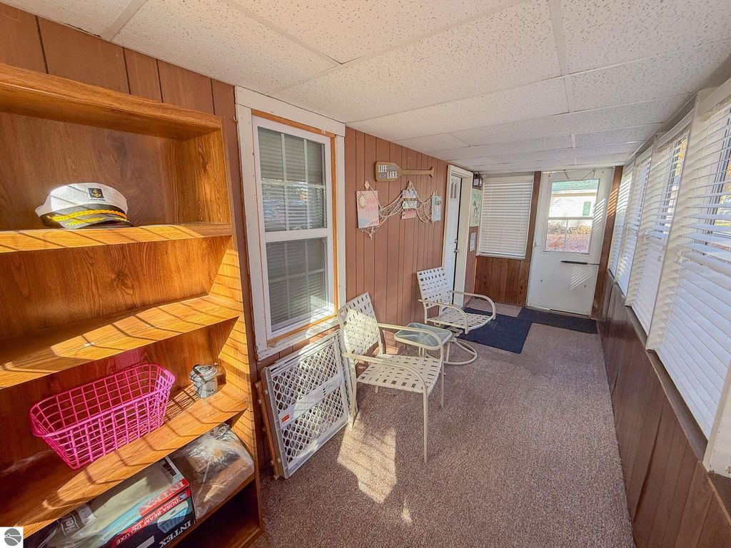 Interior view of a cozy sunroom at 133 Maple Bluffs Avenue, featuring a wooden shelf with a captain's hat, a pink basket, and two white metal chairs, emphasizing the cottage's inviting atmosphere near Houghton Lake.