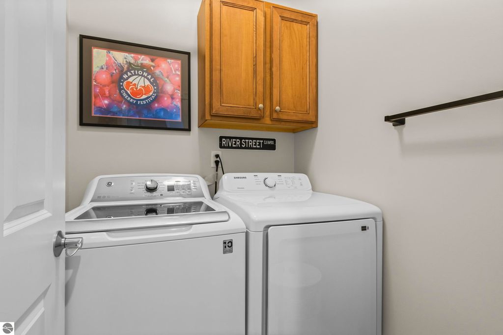 Laundry room featuring a washer and dryer, wooden cabinets, and a framed National Cherry Festival poster, emphasizing the condo's practical amenities in Traverse City.