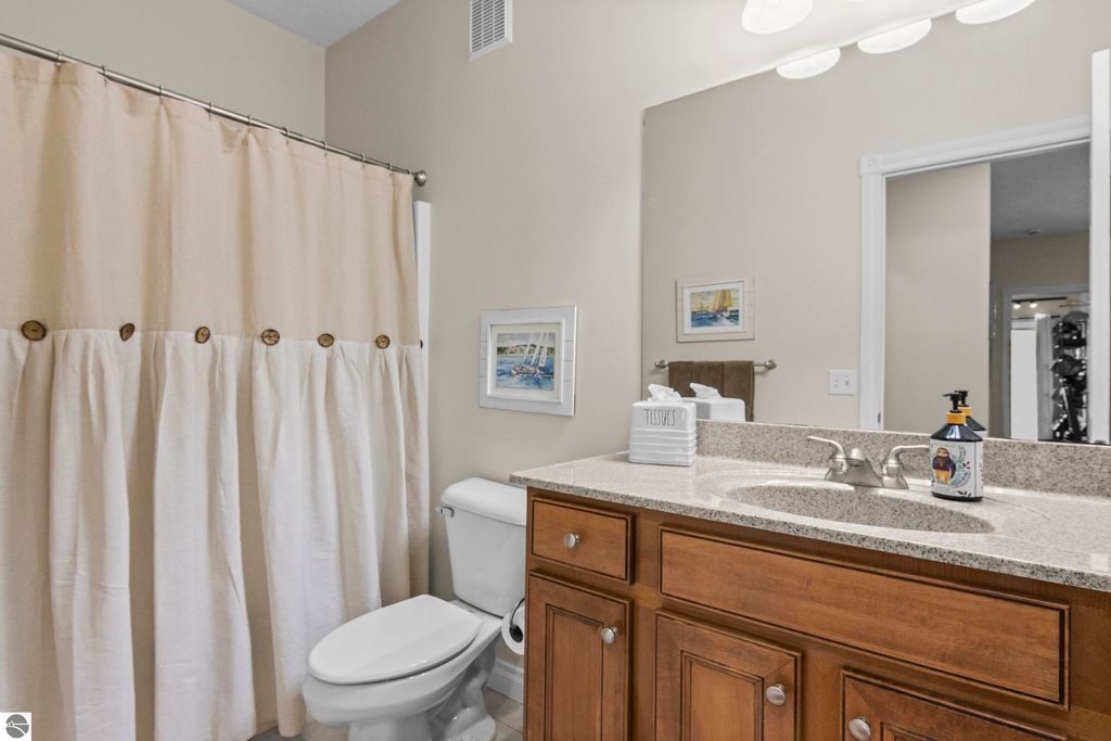 Bathroom featuring a beige shower curtain with wooden buttons, a modern vanity with wooden cabinets, a granite countertop, and a white toilet, complemented by framed artwork and decorative toiletries.