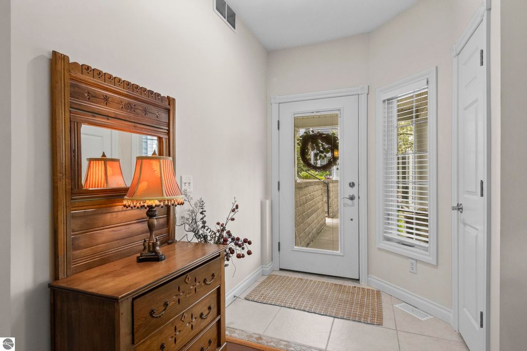 Welcoming entryway of a 2-bedroom condo featuring a wooden dresser with decorative lamp, mirror, and door leading to a private balcony, emphasizing cozy living near Traverse City.