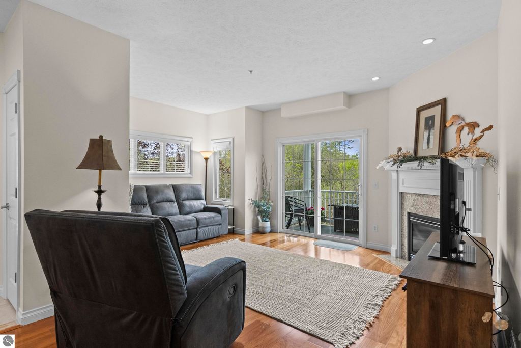 Cozy living room in a 2-bedroom condo, featuring a comfortable gray sofa, hardwood floors, a gas fireplace, and large windows with views of surrounding greenery, enhancing the serene atmosphere near Traverse City, MI.